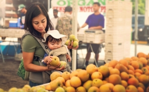 A young mom shops for fruit from organic farmers at an outdoor market. She holds her baby in front of her.