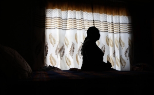 A woman, shown in shadow, sits posing in profile against the backdrop of a light white curtain with beige and brown designs.